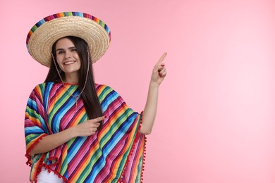 Photo of Young woman in Mexican sombrero hat and poncho pointing at something on pink background. Space for text