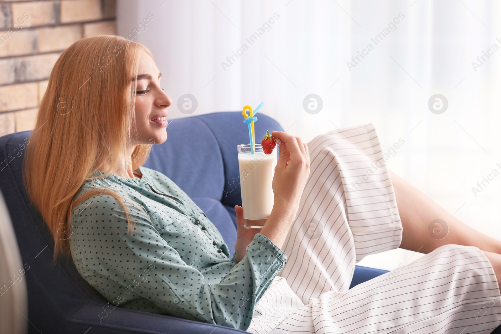 Photo of Young woman with glass of delicious milk shake in armchair indoors