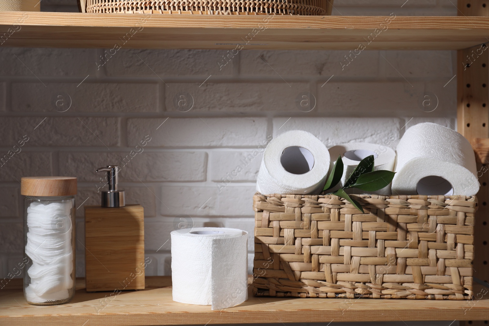 Photo of Toilet paper rolls in wicker basket, green leaves, cotton pads and dispenser on wooden shelf against white brick wall