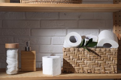 Toilet paper rolls in wicker basket, green leaves, cotton pads and dispenser on wooden shelf against white brick wall