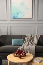 Red apples, book and glasses on wooden nesting tables near grey sofa with blanket in room