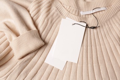 Photo of Blank white tags on beige sweater, top view. Space for text