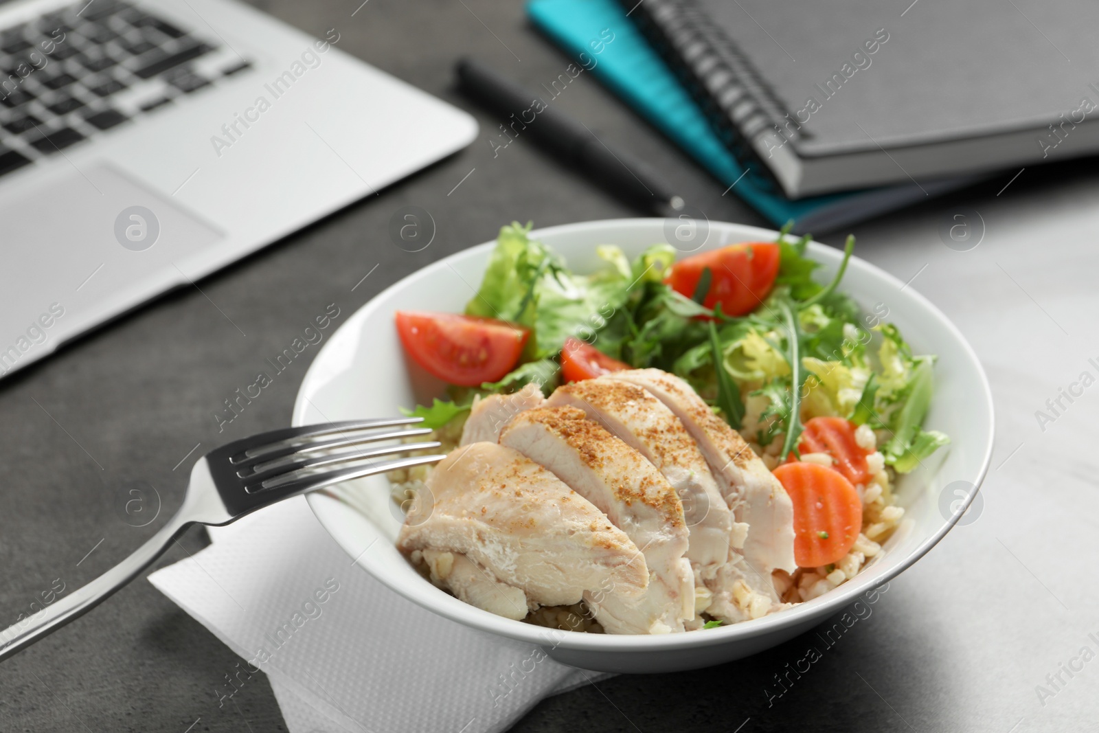 Photo of Bowl of tasty food, laptop, fork and notebooks on grey table. Business lunch