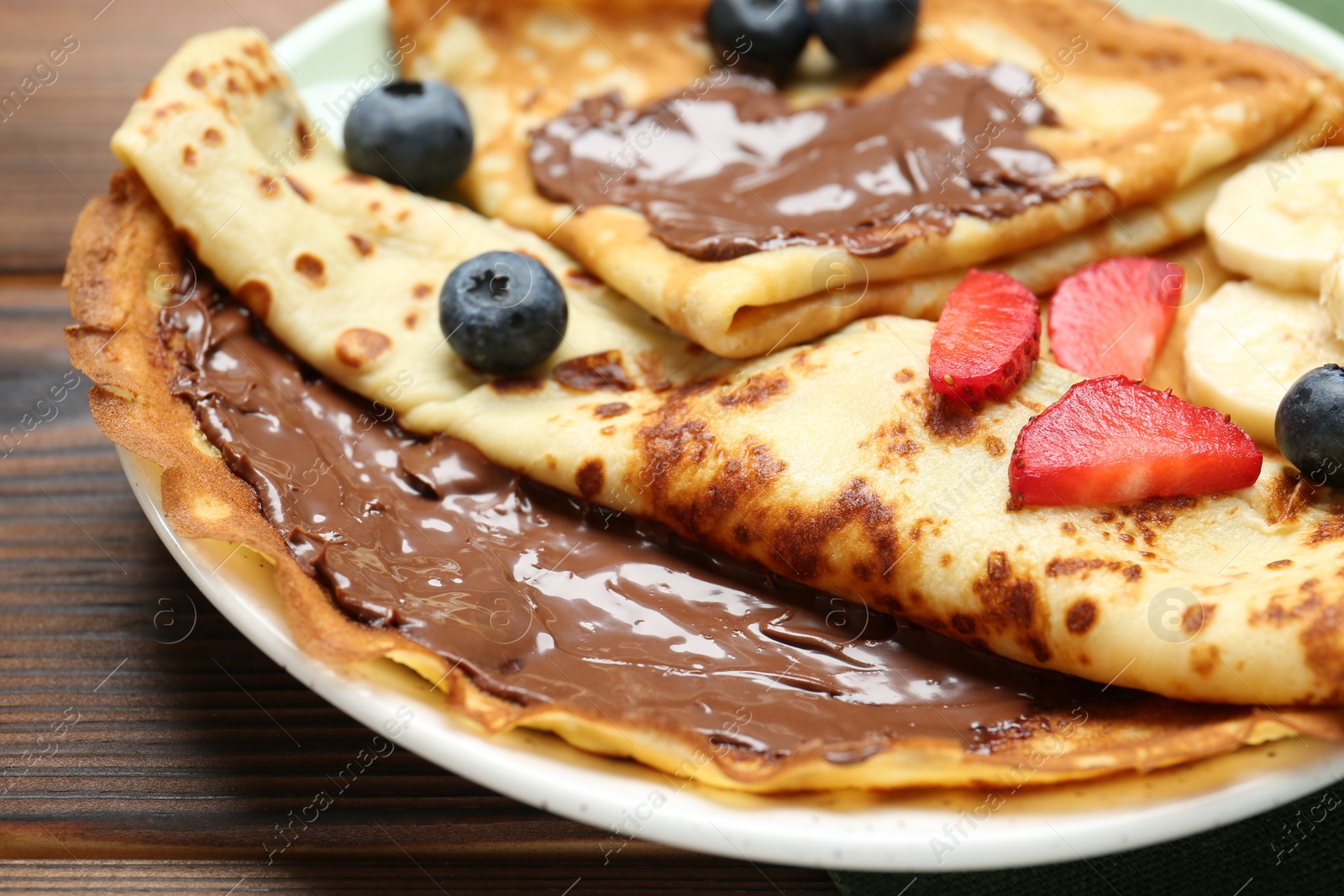Photo of Tasty crepes with chocolate paste, banana and berries on wooden table, closeup