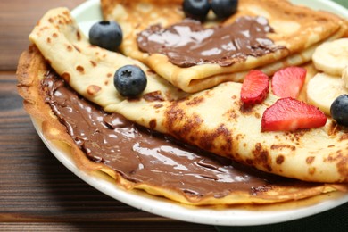 Tasty crepes with chocolate paste, banana and berries on wooden table, closeup