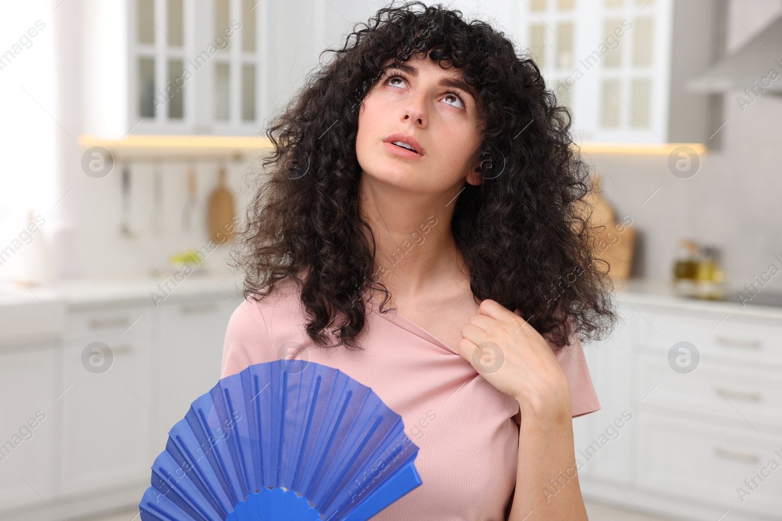 Photo of Young woman waving blue hand fan to cool herself in kitchen