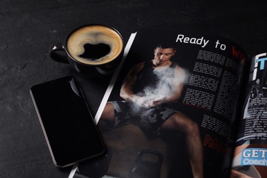 Photo of Open sports magazine, smartphone with blank screen and cup of coffee on black table, closeup