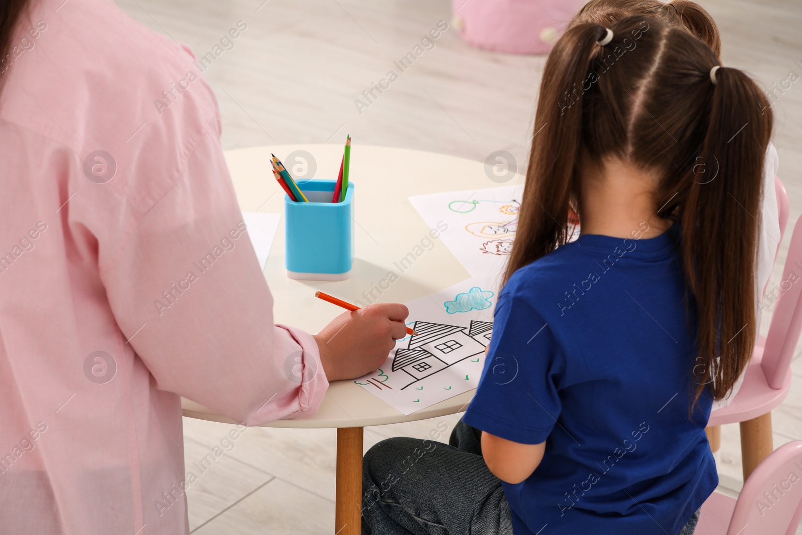 Photo of Little child and nursery teacher drawing pictures at desk in kindergarten, back view. Playtime activities