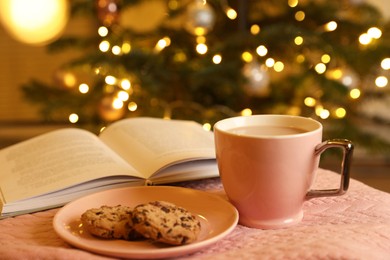 Photo of Cup of delicious cocoa, cookies and open book on pink fabric against blurred Christmas tree with lights