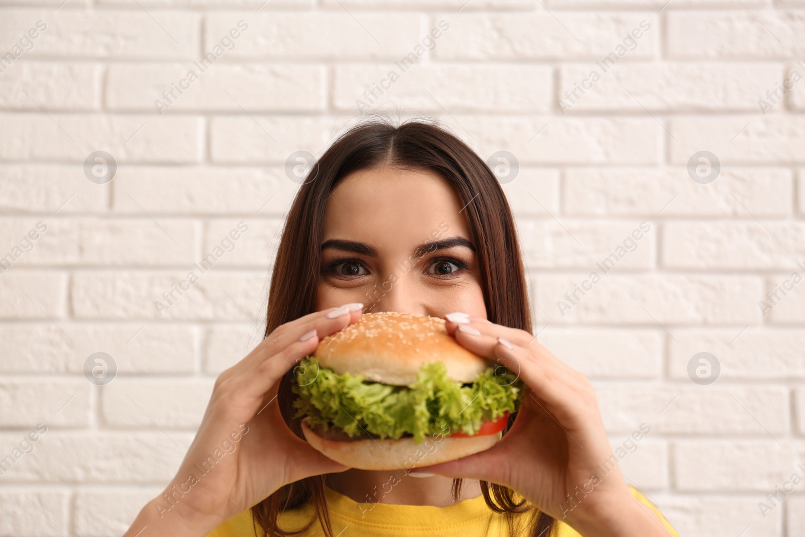 Photo of Young woman eating tasty burger near brick wall
