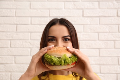 Young woman eating tasty burger near brick wall