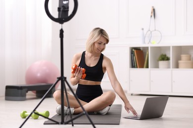 Photo of Smiling sports blogger with dumbbells streaming online fitness lesson with phone at home