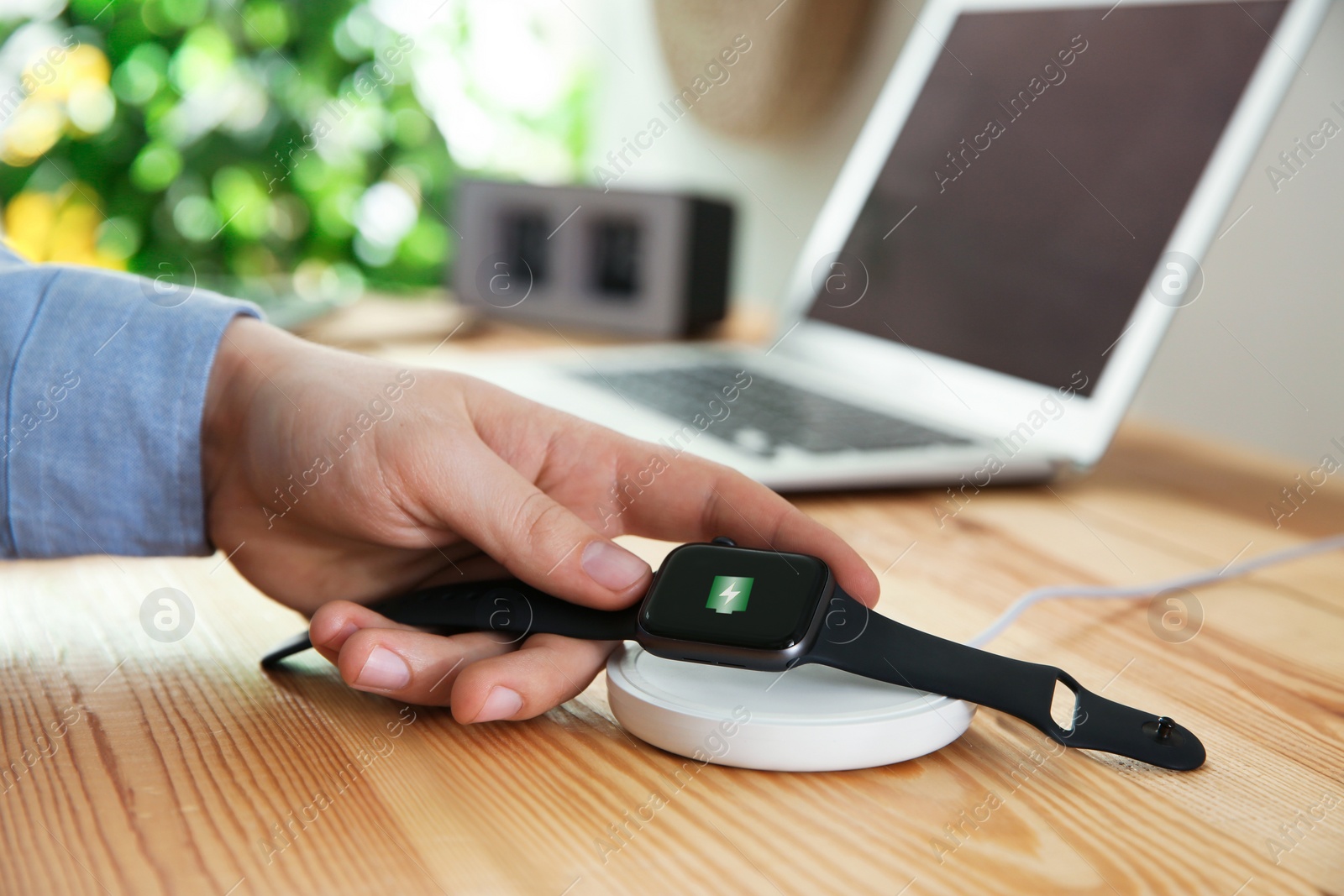 Photo of Man putting smartwatch onto wireless charger at wooden table, closeup. Modern workplace accessory