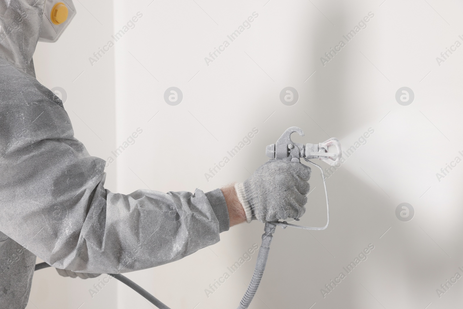 Photo of Decorator in protective overalls painting wall with spray gun indoors, closeup