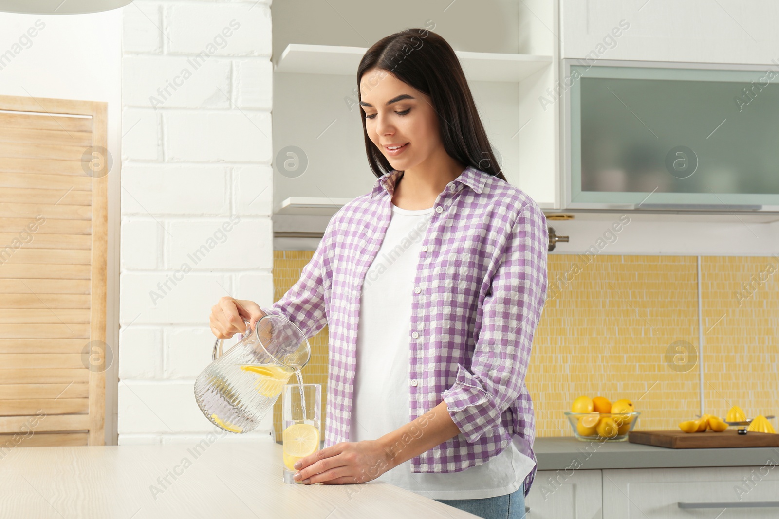 Photo of Beautiful young woman pouring lemon water into glass from jug in kitchen