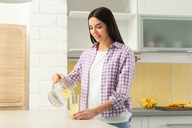 Beautiful young woman pouring lemon water into glass from jug in kitchen