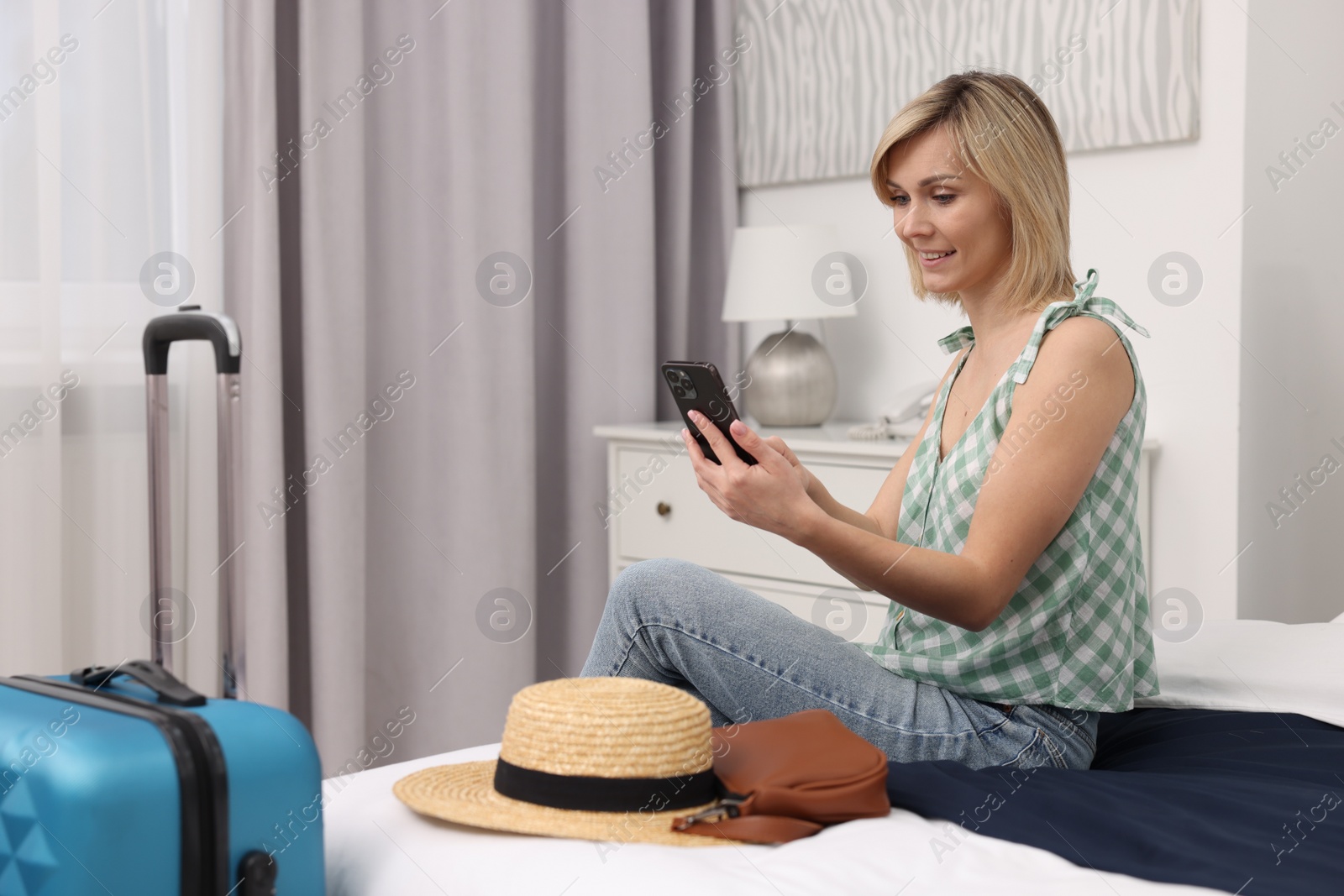 Photo of Smiling guest with smartphone relaxing on bed in stylish hotel room