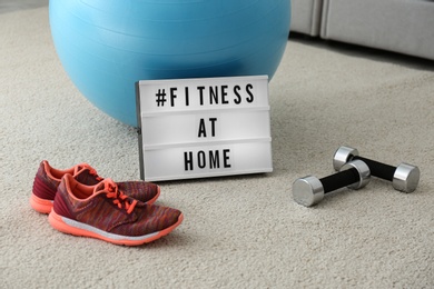 Photo of Sport equipment and lightbox with hashtag FITNESS AT HOME on floor indoors. Message to promote self-isolation during COVID‑19 pandemic