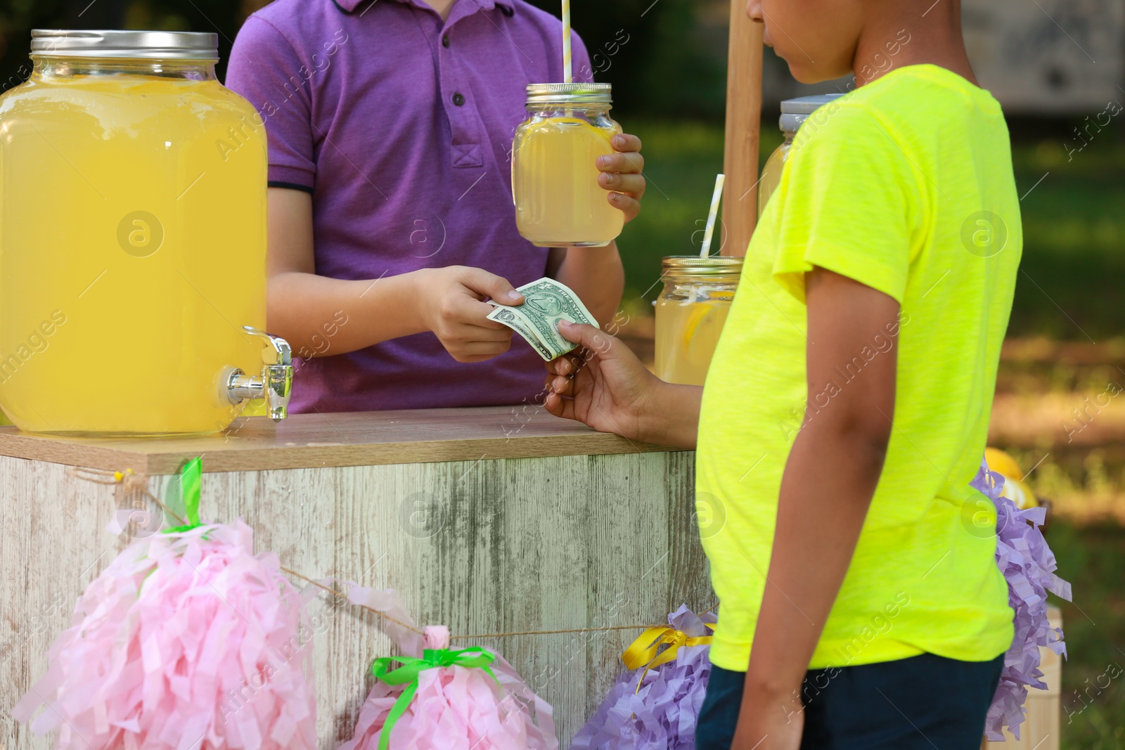 Photo of Little boy selling natural lemonade to African-American kid in park, closeup. Summer refreshing drink