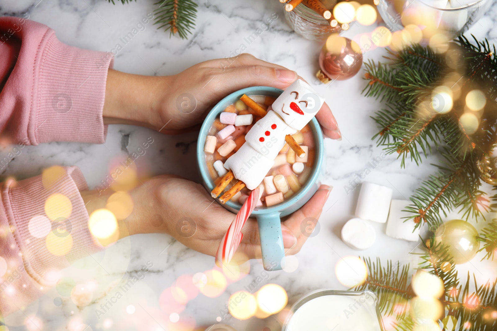 Image of Woman holding cup with hot drink and marshmallow snowman at white marble table, top view. Bokeh effect