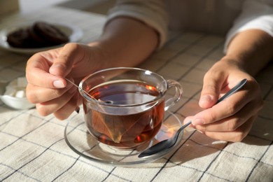Photo of Woman holding spoon near glass cup with tea bag at table, closeup