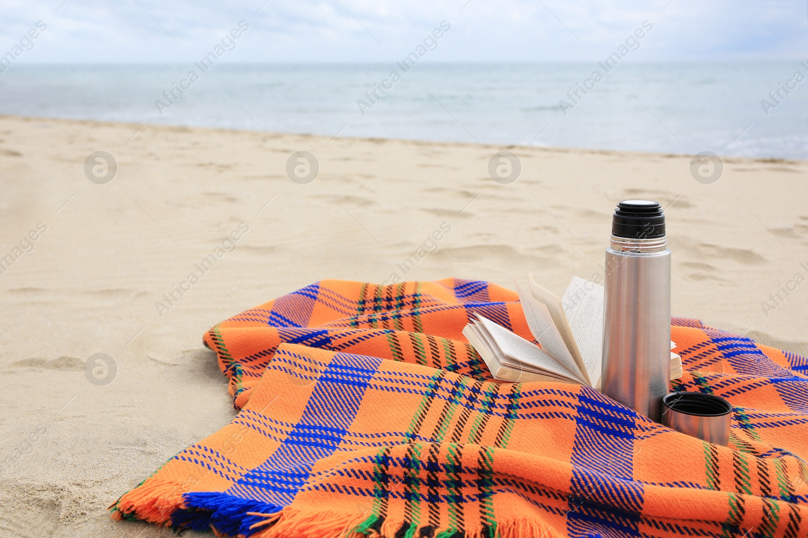 Photo of Metallic thermos with hot drink, open book and plaid on sandy beach near sea, space for text