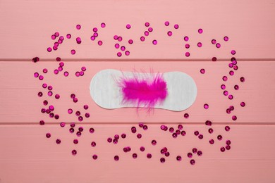 Photo of Sanitary pad with feather surrounded by sequins on pink wooden background, flat lay. Menstrual cycle