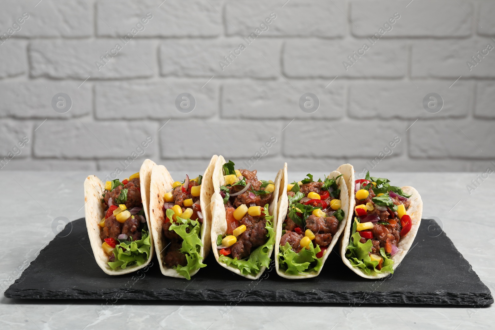 Photo of Delicious tacos with meat and vegetables on light gray marble table against brick wall