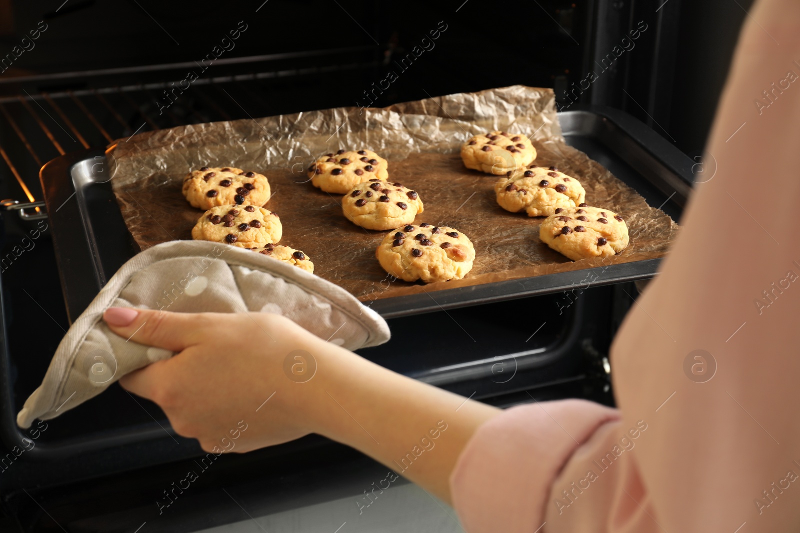 Photo of Woman taking out delicious chocolate chip cookies from oven, closeup