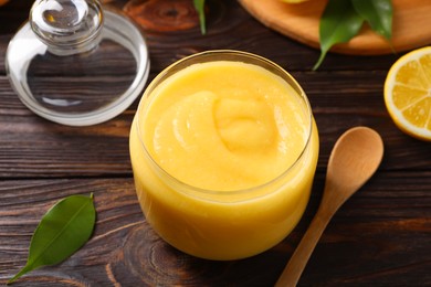 Photo of Delicious lemon curd in glass jar, fresh citrus fruit and spoon on wooden table