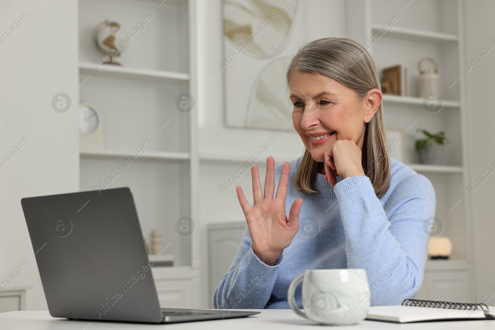 Photo of Happy woman waving hello during video call at table indoors