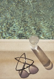 Stylish sunglasses and glass of water near outdoor swimming pool on sunny day, above view