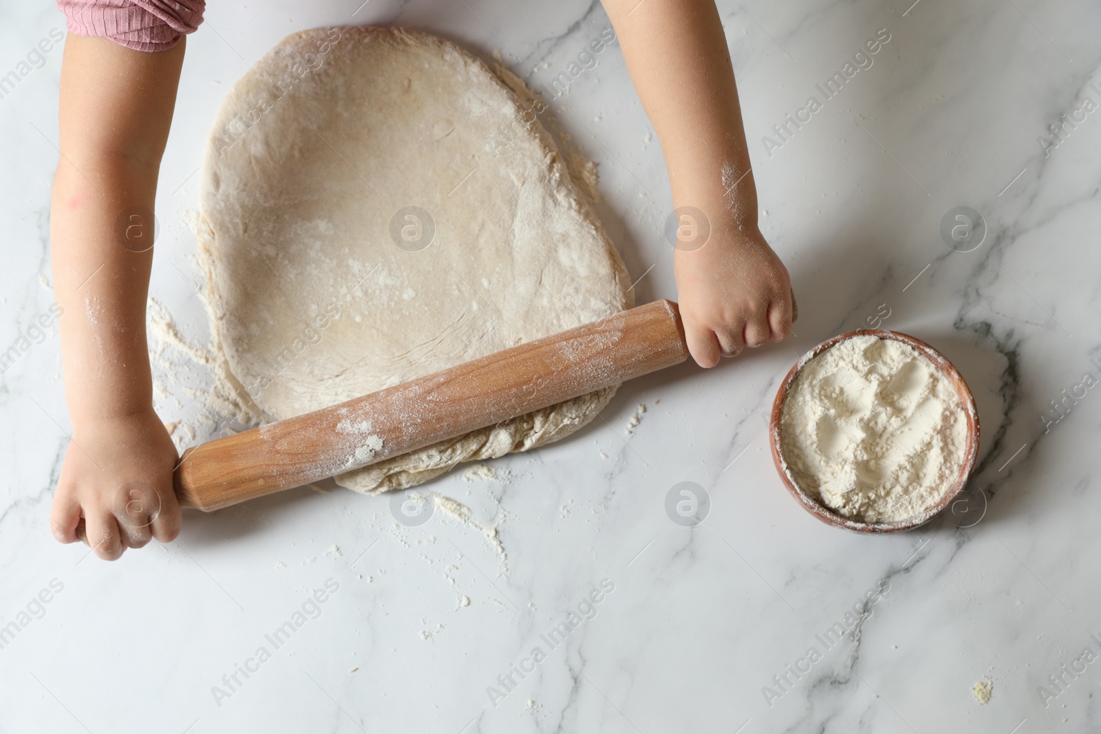 Photo of Little child rolling raw dough at white table, top view