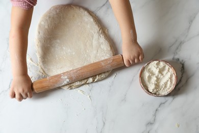 Little child rolling raw dough at white table, top view