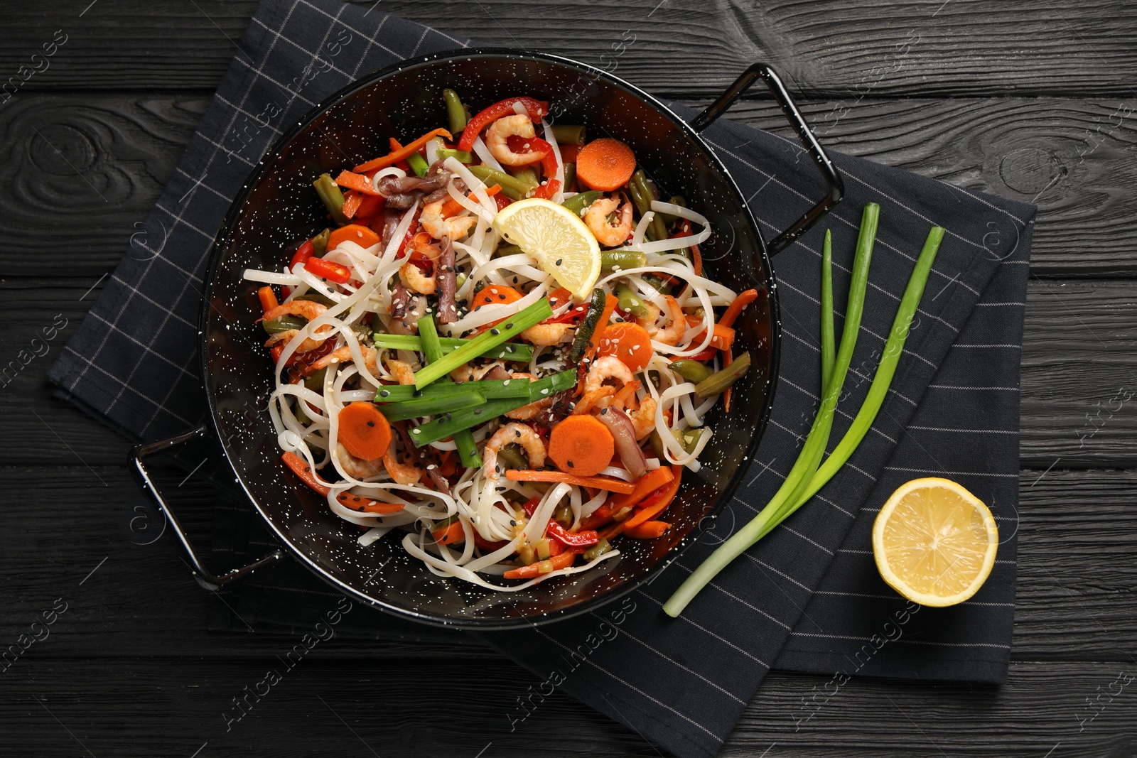 Photo of Shrimp stir fry with noodles and vegetables in wok on black wooden table, top view