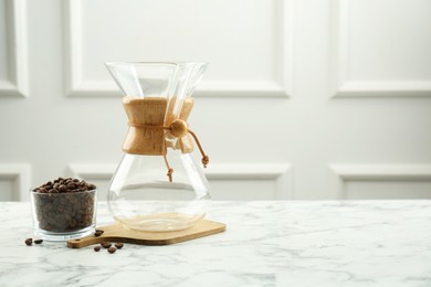 Photo of Empty glass chemex coffeemaker and beans in bowl on white marble table, space for text