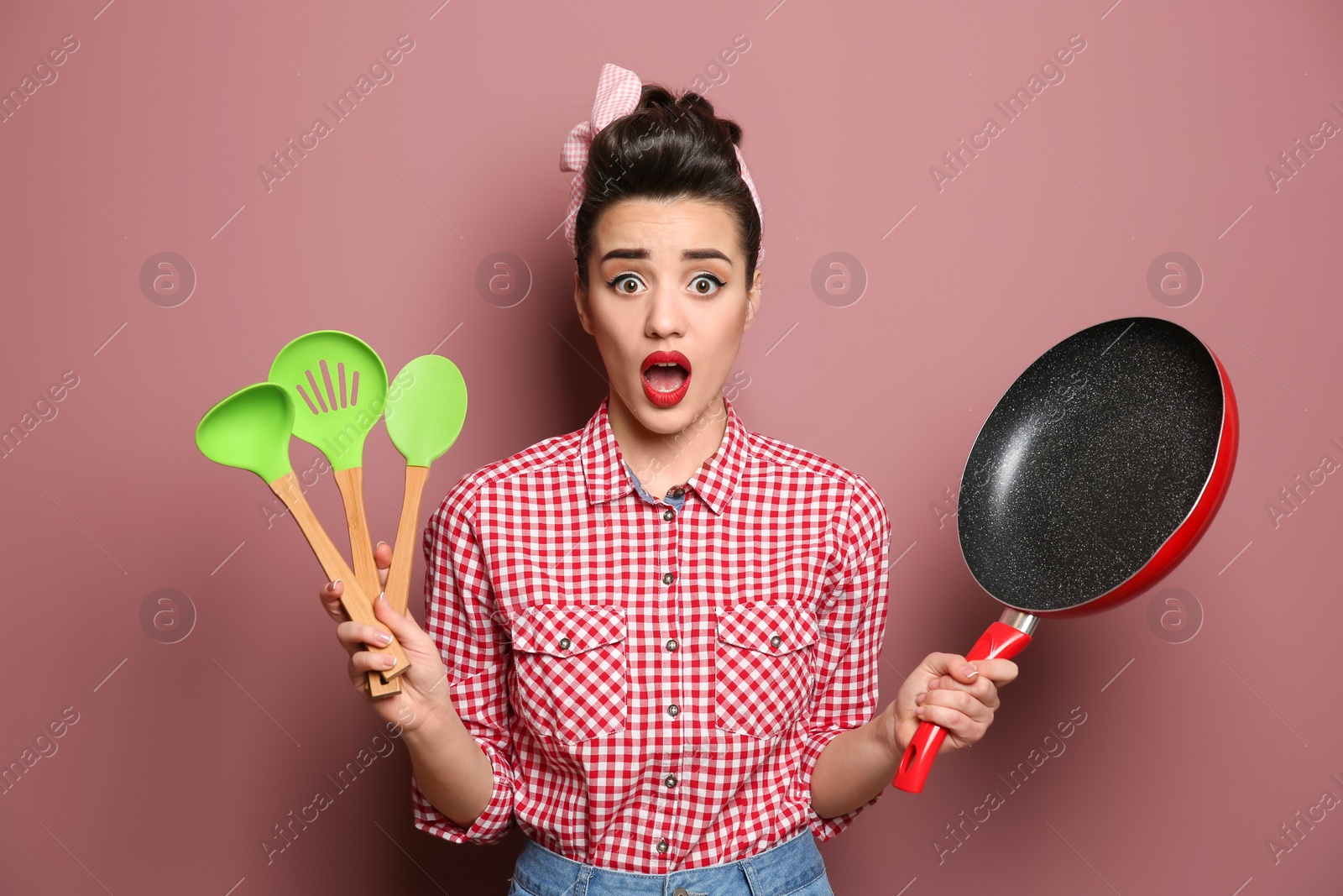 Photo of Funny young housewife with frying pan and cooking utensils on color background