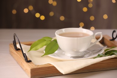 Photo of Cup of aromatic tea with milk, spoon, saucer and green leaves on light wooden table