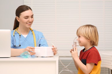 Photo of Medical assistant sitting near sick little boy while he using nebulizer for inhalation in hospital