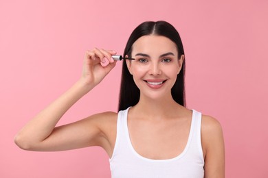 Beautiful woman applying serum onto her eyelashes on pink background. Cosmetic product