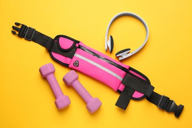 Photo of Stylish pink waist bag, dumbbells and headphones on yellow background, flat lay