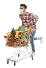 Young man in medical mask with shopping cart full of groceries on white background