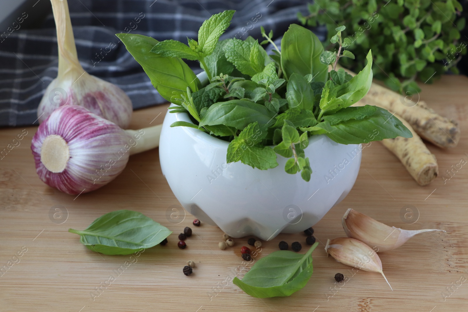 Photo of Mortar with different fresh herbs near garlic, horseradish roots and black peppercorns on wooden table, closeup