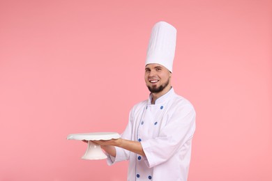 Photo of Happy professional confectioner in uniform holding empty cake stand on pink background. Space for text