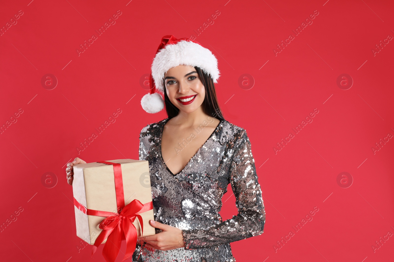 Photo of Woman in silver dress and Santa hat holding Christmas gift on red background