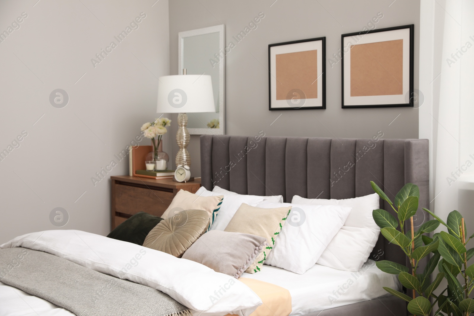 Photo of Comfortable bed with cushions, lamp and different decor on wooden chest of drawers in room. Stylish interior