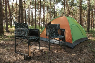 Photo of Camouflage chairs with hat and axe near camping tent in forest on sunny day