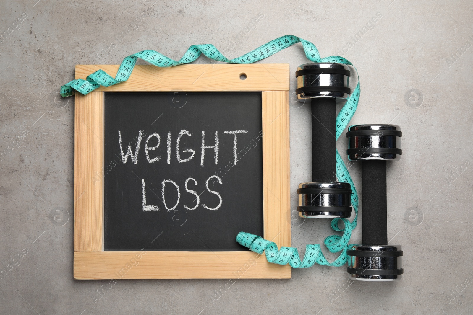 Photo of Small chalkboard with phrase Weight Loss, dumbbells and measuring tape on grey table, flat lay
