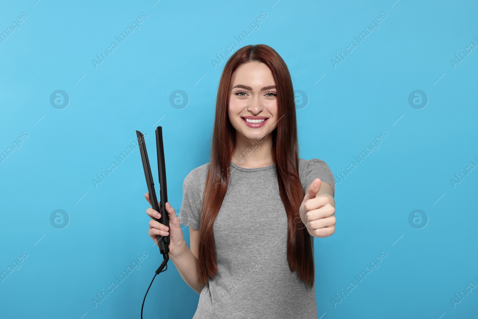 Photo of Beautiful woman with hair iron showing thumbs up on light blue background