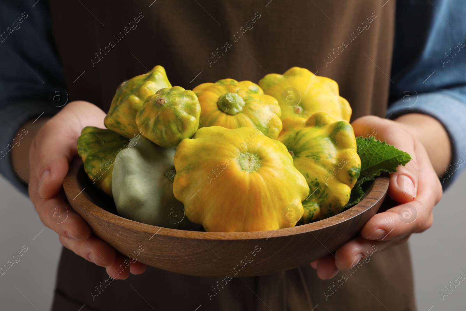 Photo of Woman with wooden bowl of fresh ripe pattypan squashes on grey background, closeup